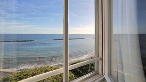 Vista costera desde una ventana, con el mar, un rompeolas rocoso y una playa.
