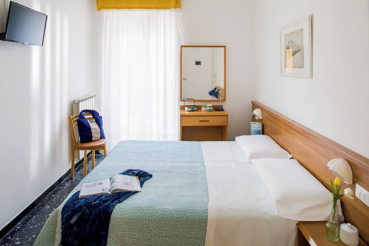 Een hotelkamer met een bed, nachtkastje, spiegel, een aan de muur bevestigde televisie en een raam met gordijnen.