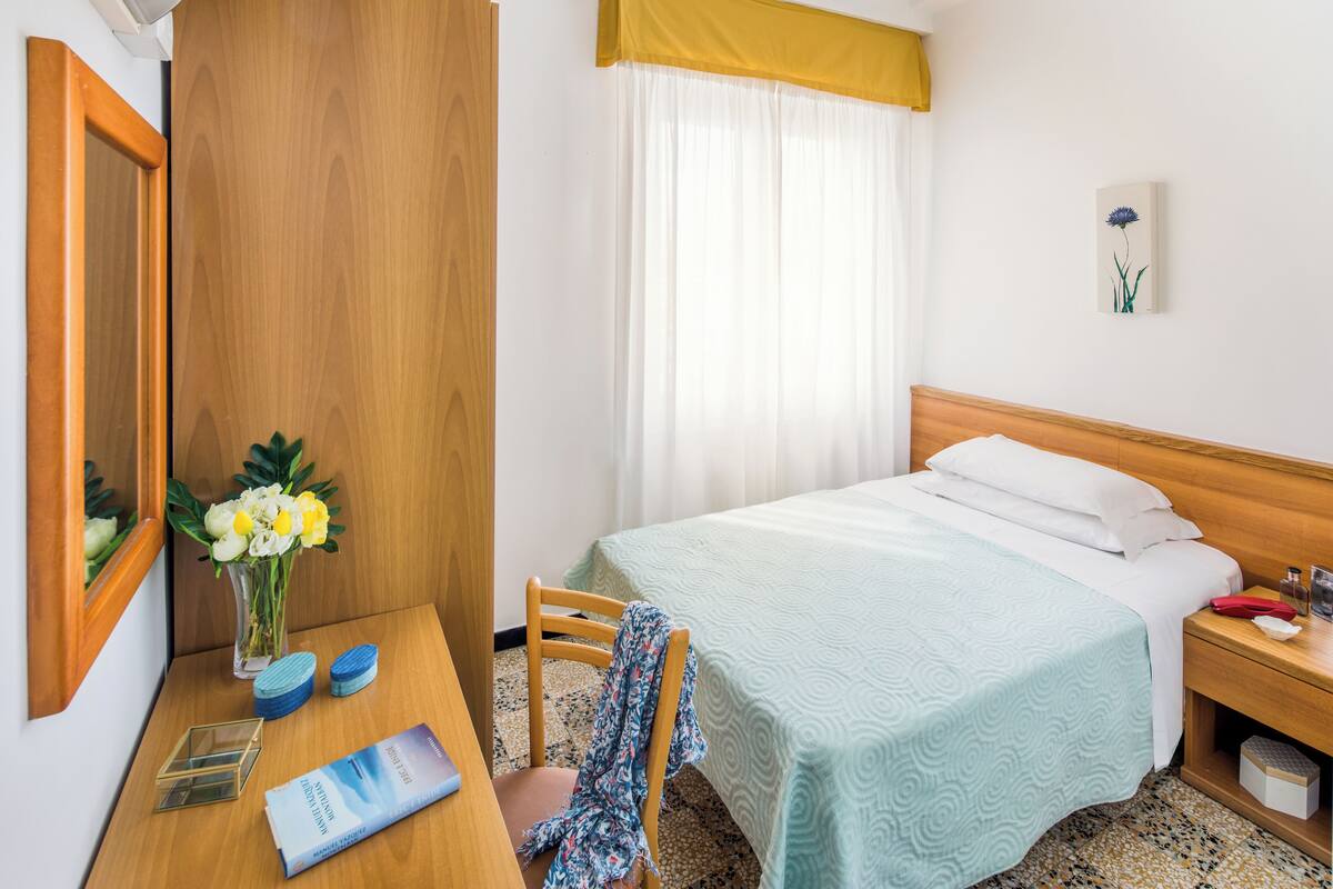 Een hotelkamer met een bed, een houten nachtkastje, een bureau met stoel, een spiegel, een vaas met bloemen, en een raam met gordijnen.