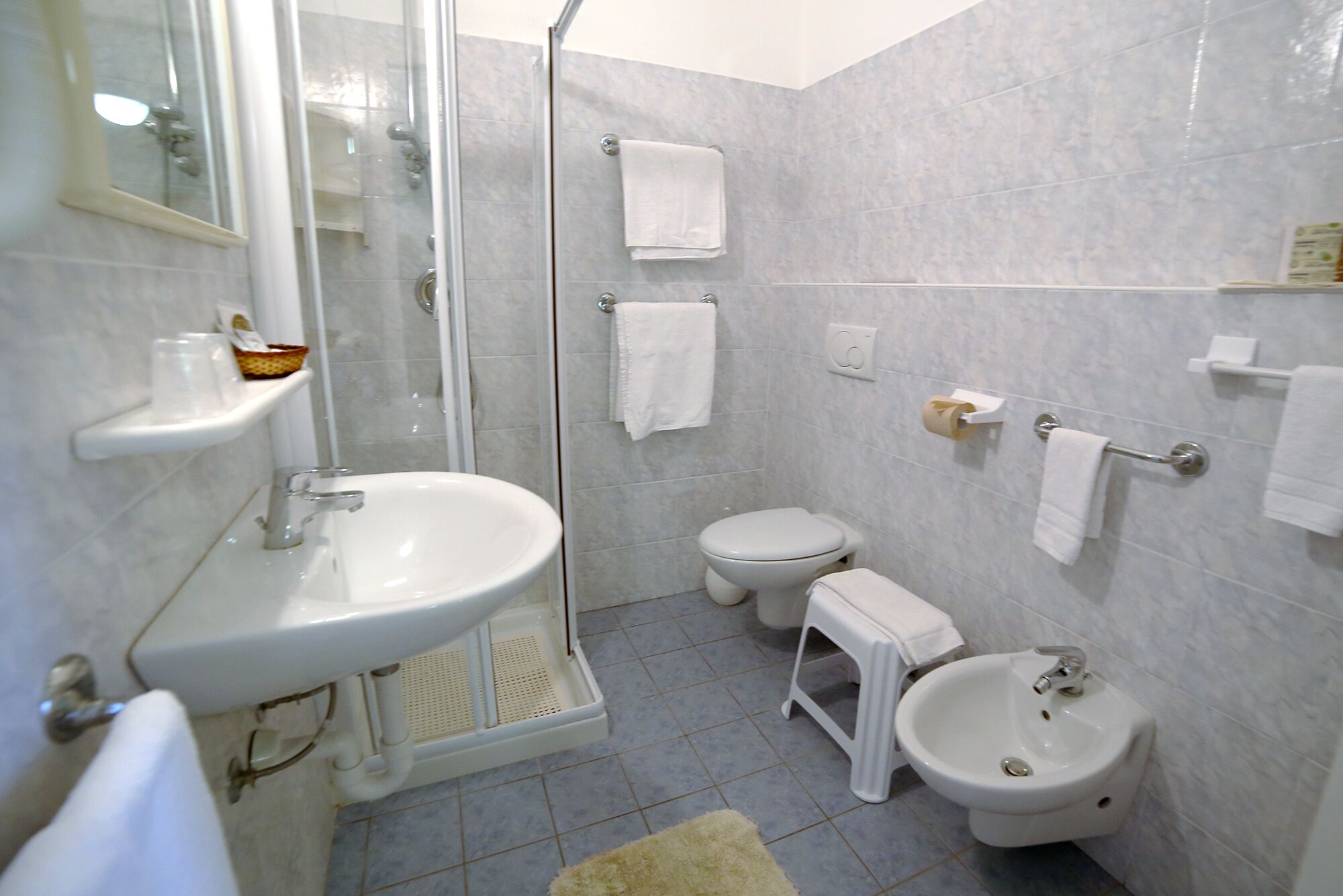 A bathroom with a glass-enclosed shower, a sink, a toilet, a bidet, and a small stool.