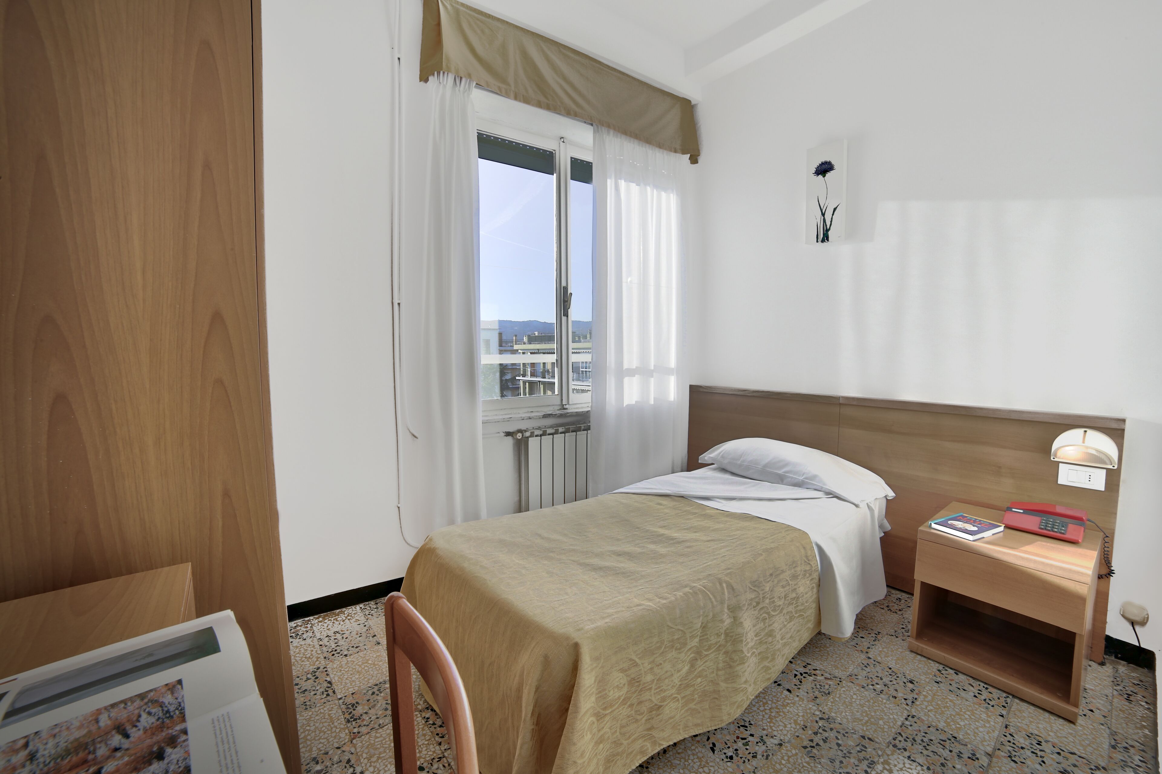 A hotel room with a bed, a nightstand, a chair, a window with curtains, and a view of buildings outside.