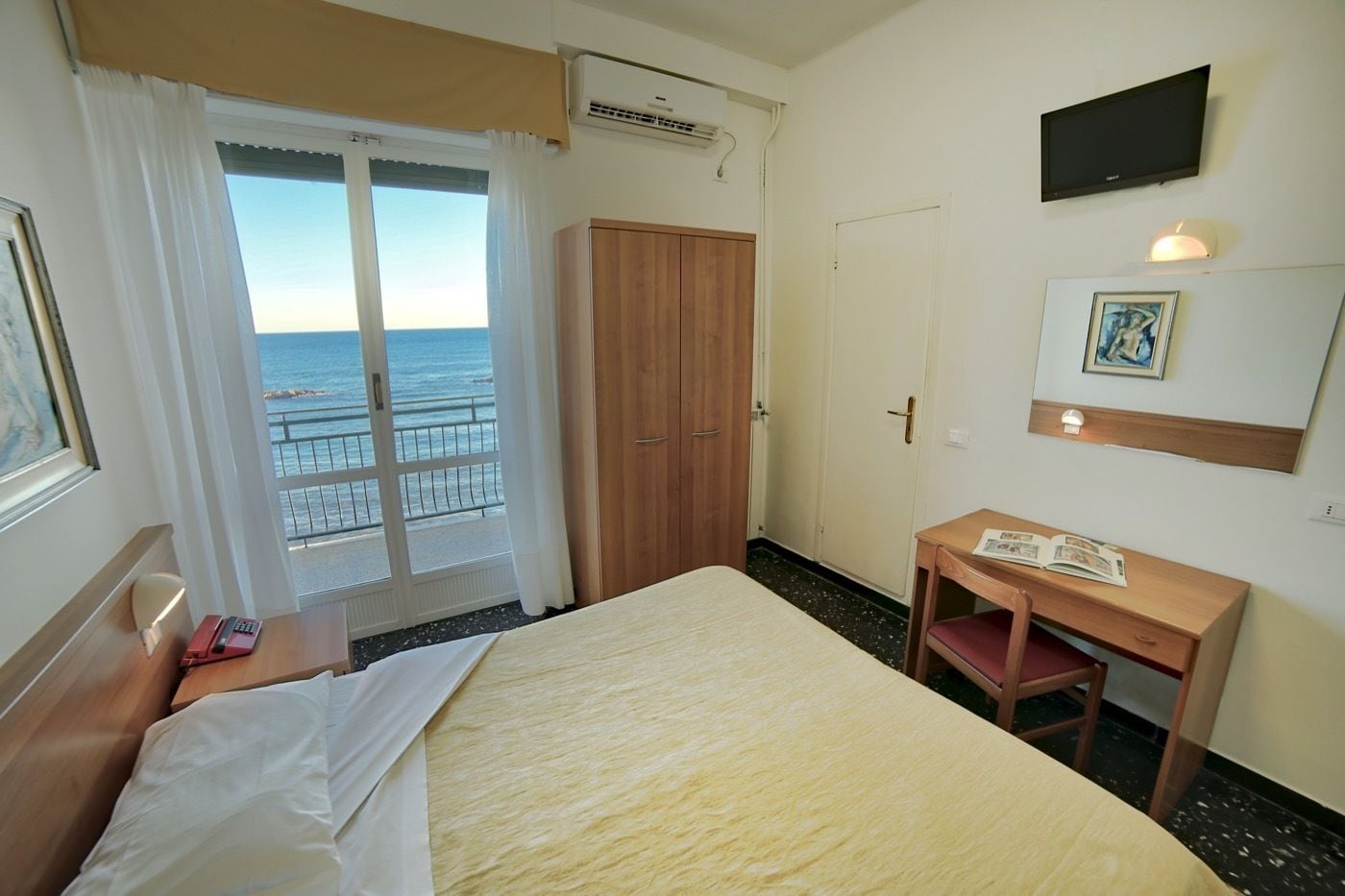 Ein Hotelzimmer mit Bett, Schreibtisch, Kleiderschrank, Fernseher und einem Fenster mit Meerblick.