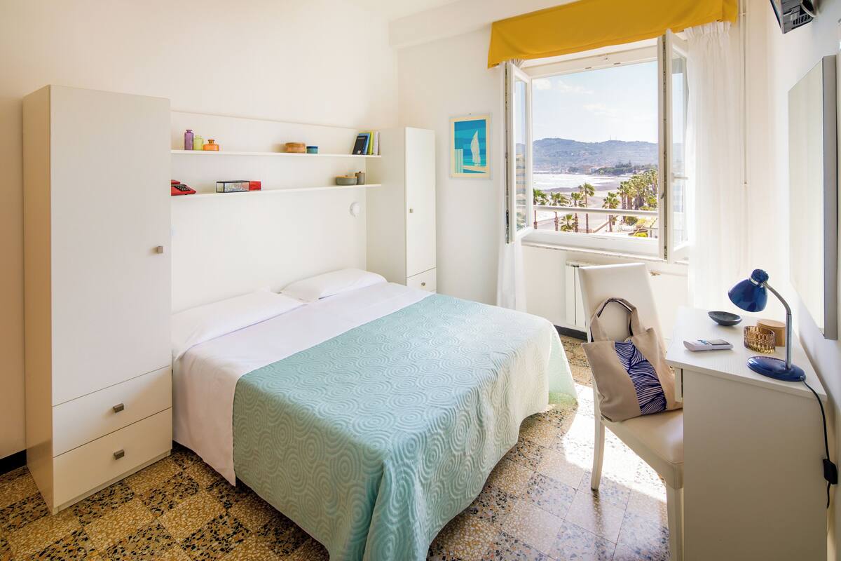 Een slaapkamer met een bed, een bureau met stoel, een raam met uitzicht op het strand en een kledingkast.