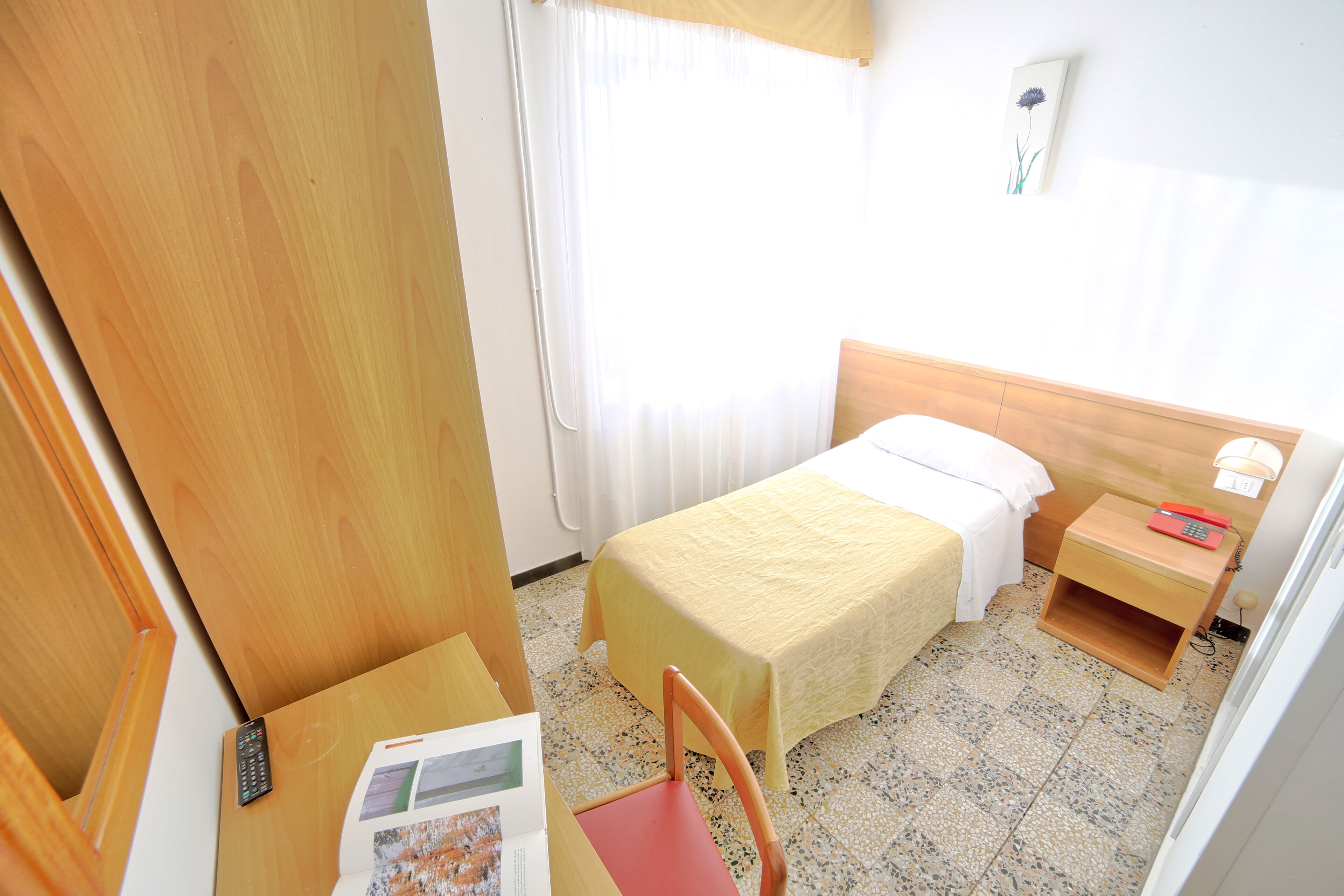 Una habitación pequeña y luminosa con una cama, mesita de noche, silla y escritorio, junto con un mando a distancia y un libro.