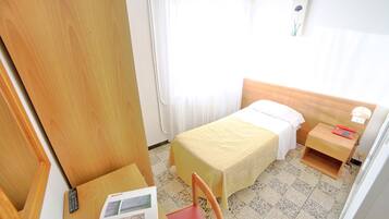 Una habitación pequeña y luminosa con una cama, mesita de noche, silla y escritorio, junto con un mando a distancia y un libro.