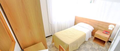 Quarto pequeno e bem iluminado com cama, mesa de cabeceira, cadeira e escrivaninha com controle remoto e um livro.