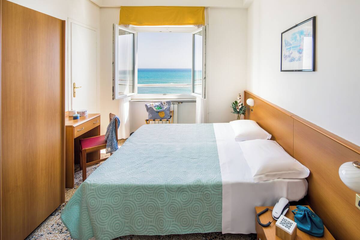 Een hotelkamer met een bed, een bureau, een stoel, een raam met uitzicht op zee en een ingelijst schilderij aan de muur.