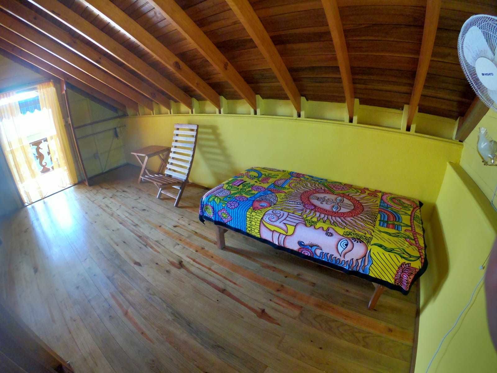 Un dormitorio ático pequeño y con una sola habitación, con vigas de madera, una cama con colcha estampada de colores, una silla y un ventilador en la pared.