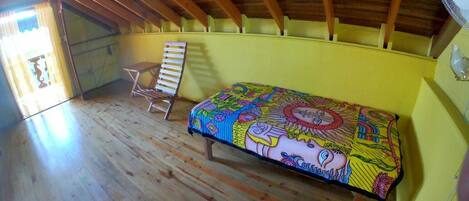 Un dormitorio ático pequeño y con una sola habitación, con vigas de madera, una cama con colcha estampada de colores, una silla y un ventilador en la pared.