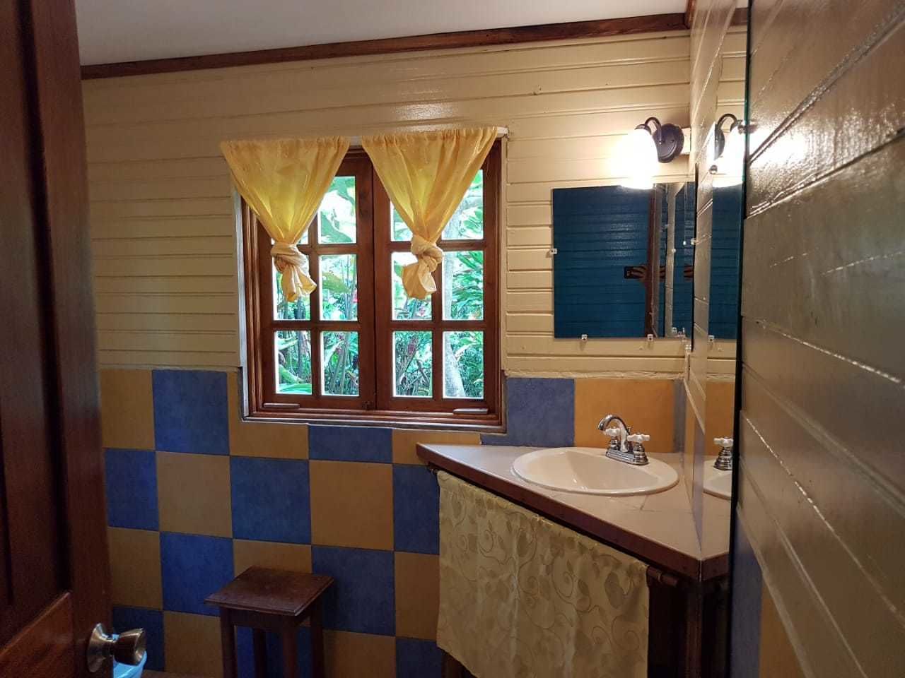 A bathroom with a checkered tile wall, a window with yellow curtains, a sink, and a mirror.