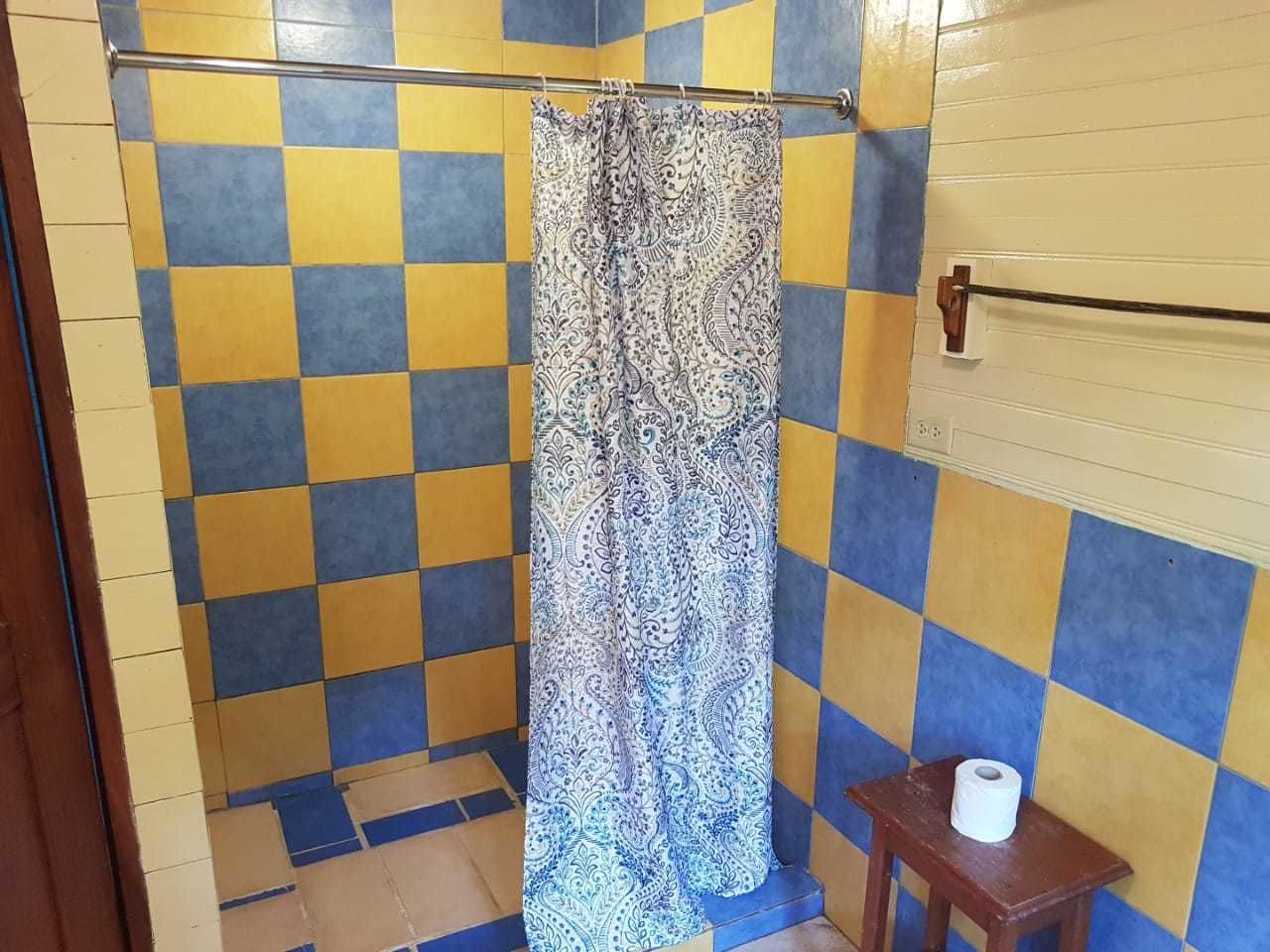 Un baño con baldosas amarillas y azules a cuadros, una cortina de ducha estampada y un taburete de madera con un rollo de papel higiénico.