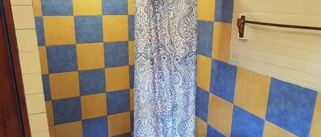 A bathroom with checkered yellow and blue tiles, a patterned shower curtain, and a small wooden stool with a roll of toilet paper.