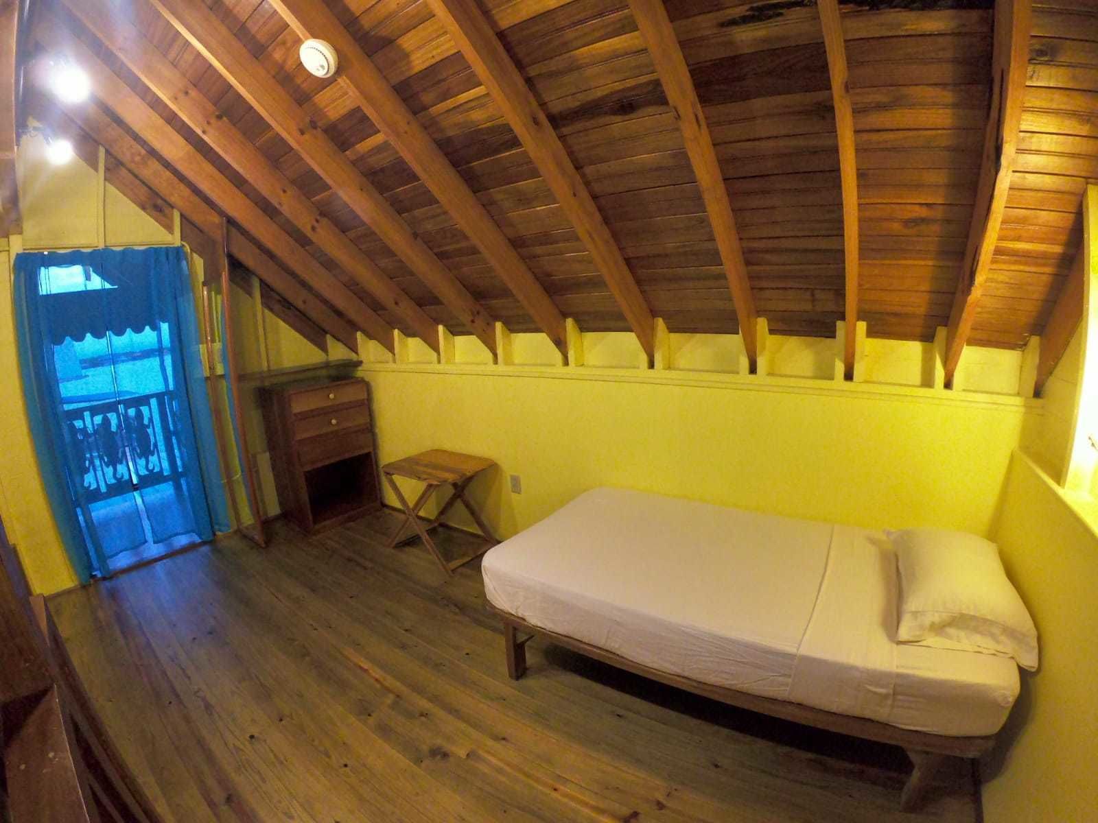 Un dormitorio ático pequeño y con una sola habitación, techo de madera, una cama, un escritorio pequeño y una ventana con cortinas azules.