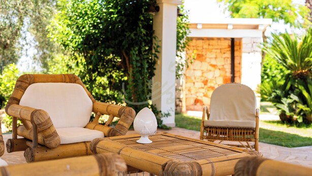 A wicker lounge chair with a cushion, a small table with a decorative item, and a stone wall in the background. - TD Villa Luna Trulli with Large Pool (Castellana Grotte)