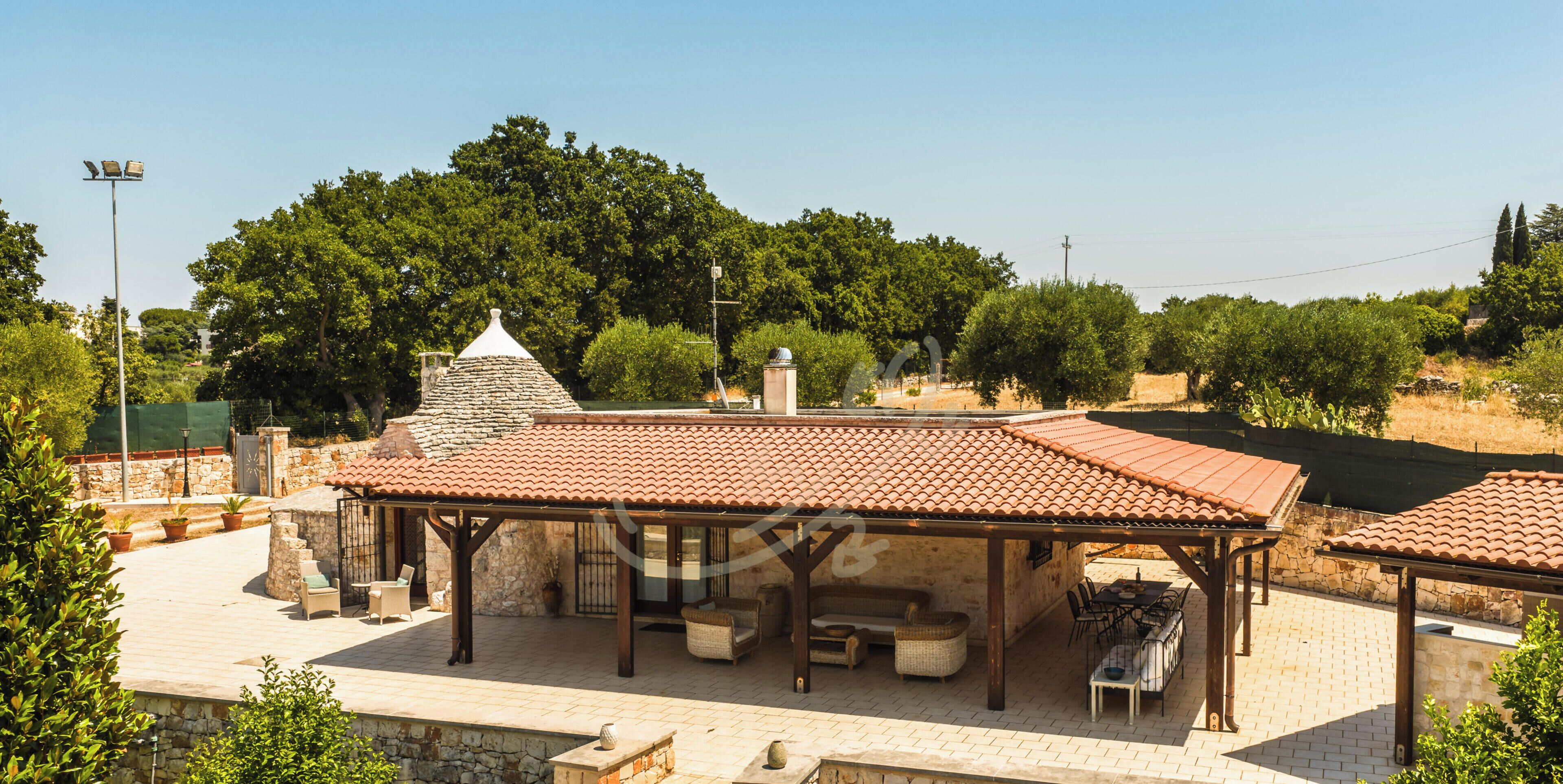 A covered outdoor seating area with stone walls and a tiled roof, surrounded by greenery and trees.