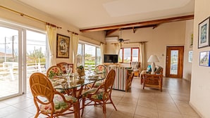 A dining area with a glass table and wicker chairs, a kitchen with a TV, and a view of the outdoors. - Villa Amarc: Family Fun, Kite Surf, Balcony View minutes to Rodgers/Baby Beach! (San Nicolas)