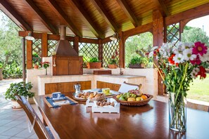 A wooden outdoor kitchen with a chimney, surrounded by potted plants and a dining table set with food and drinks. - Villa Mangini (Putignano)