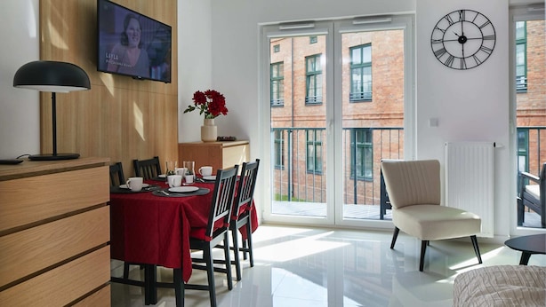 A modern living room with a dining area, a TV mounted on the wall, a clock, and a view of a brick building through the glass doors. - Praga Elegant Studio (Warsaw)