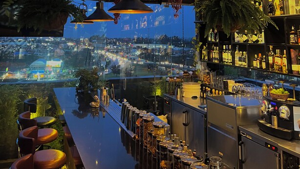 A bar area with a view of the city at dusk, featuring hanging lights and indoor plants. - Hotel Sabana Park  (Cajicá)