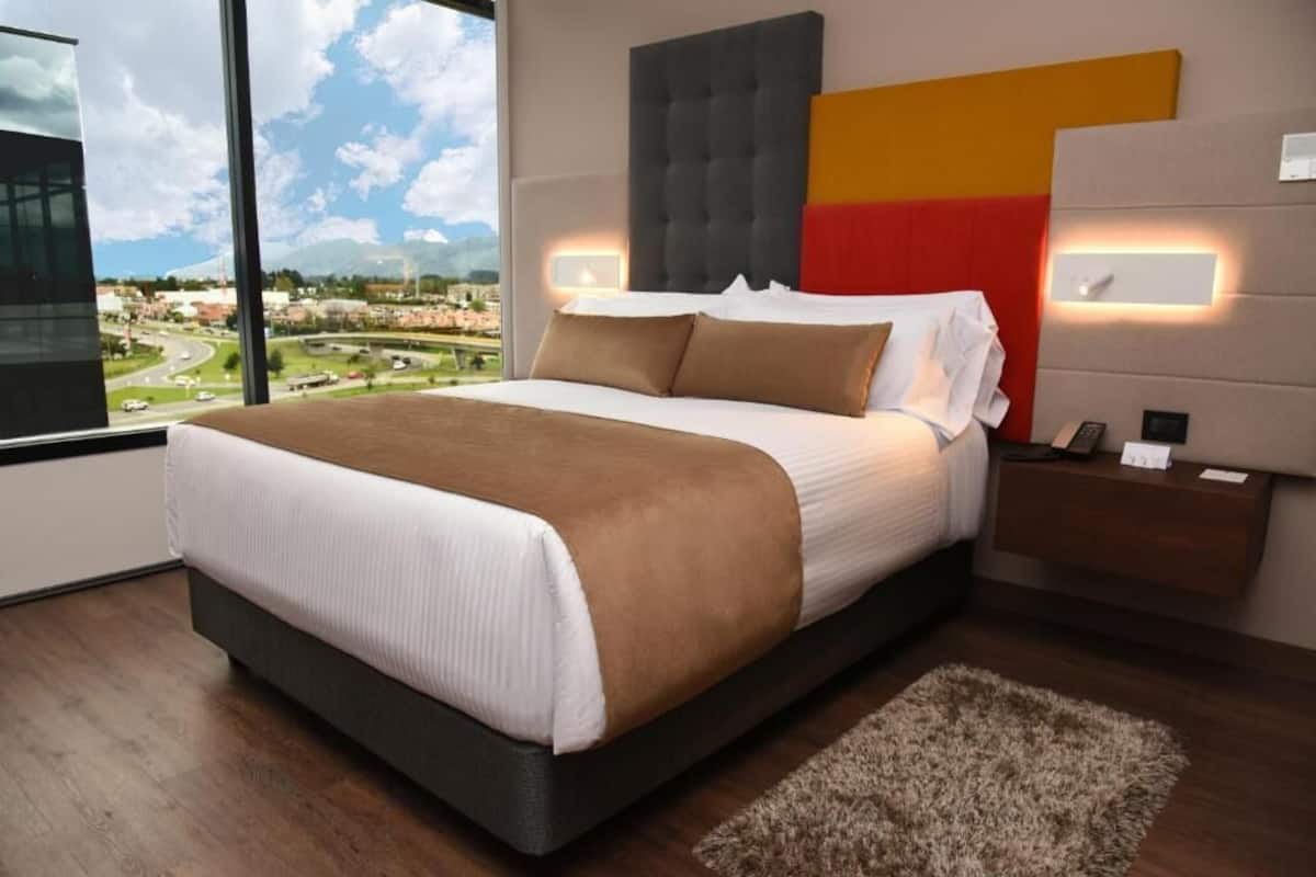 Un dormitorio moderno con una cama grande, vistas a un paisaje urbano y una vista de la ciudad a través de amplios ventanales.