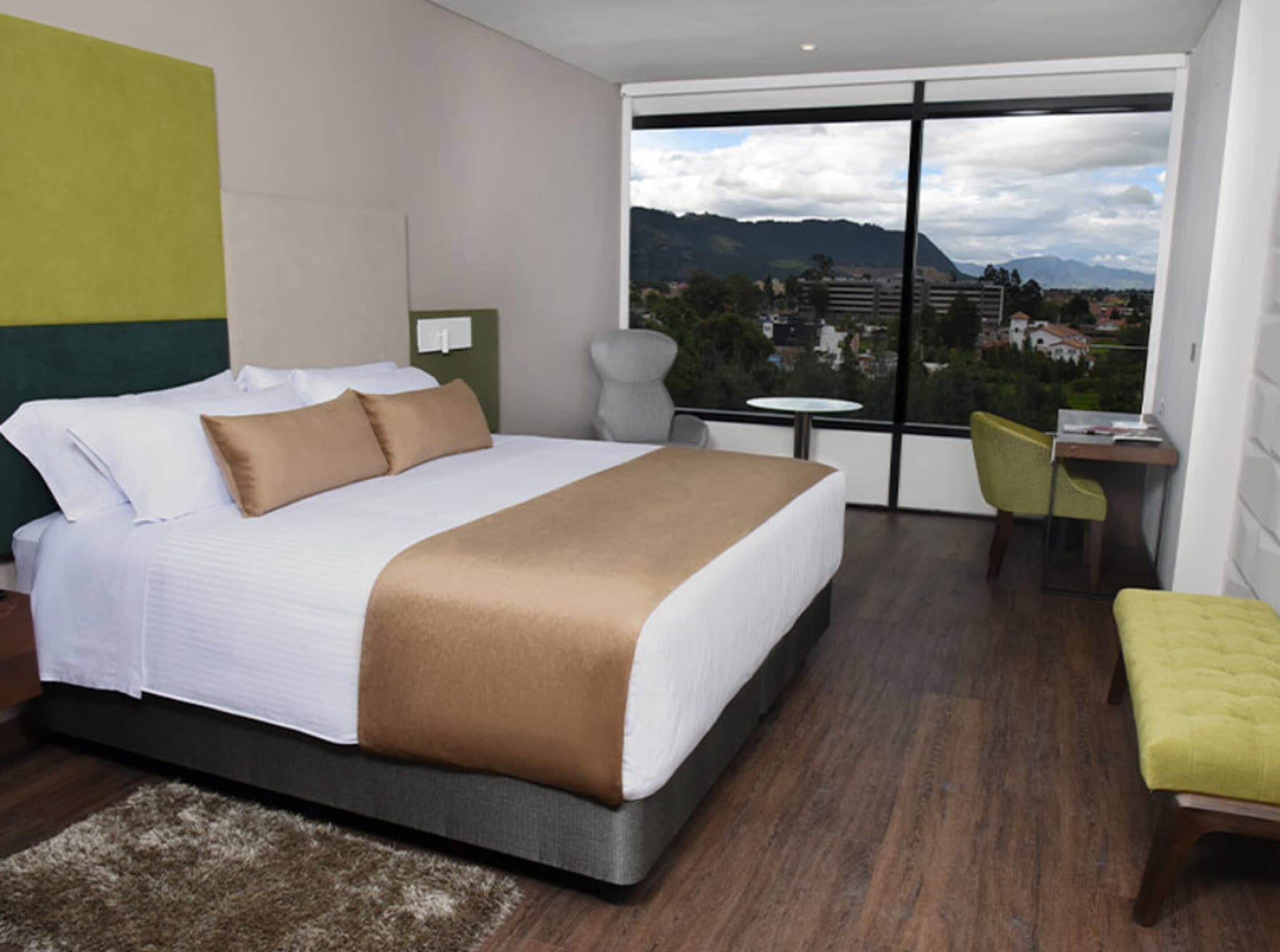Una habitación de hotel moderna con una cama grande, una mesita, una silla y vistas a un paisaje urbano y montañas.