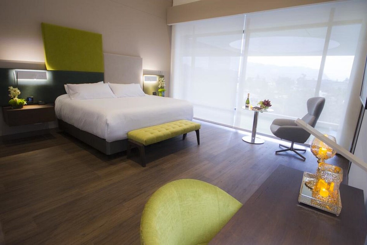 A modern hotel room with a large bed, a green armchair, a small table with a decorative item, and a large window offering a city view.