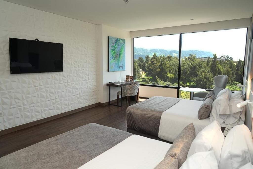 Una habitación de hotel moderna con dos camas, un escritorio, una silla, un televisor y un amplio ventanal con vistas a la montaña.