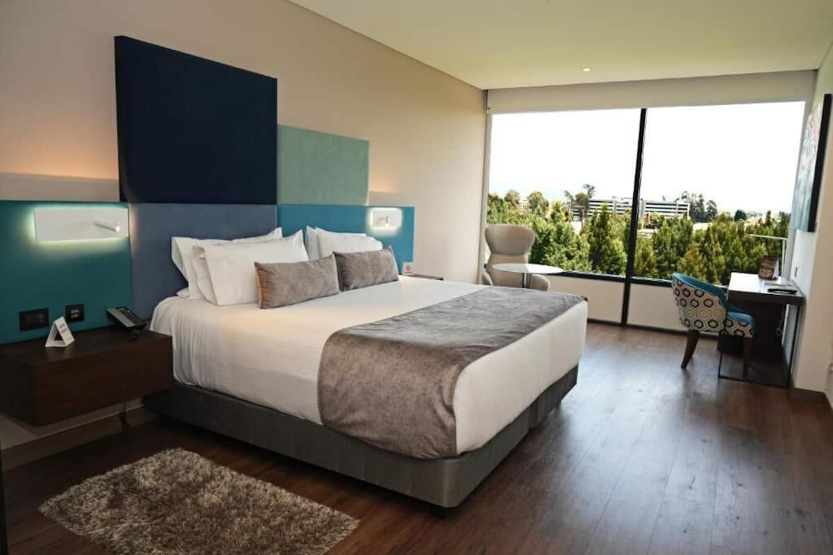 Un dormitorio moderno con una cama grande, un escritorio y vistas a la vegetación a través de amplias ventanas.