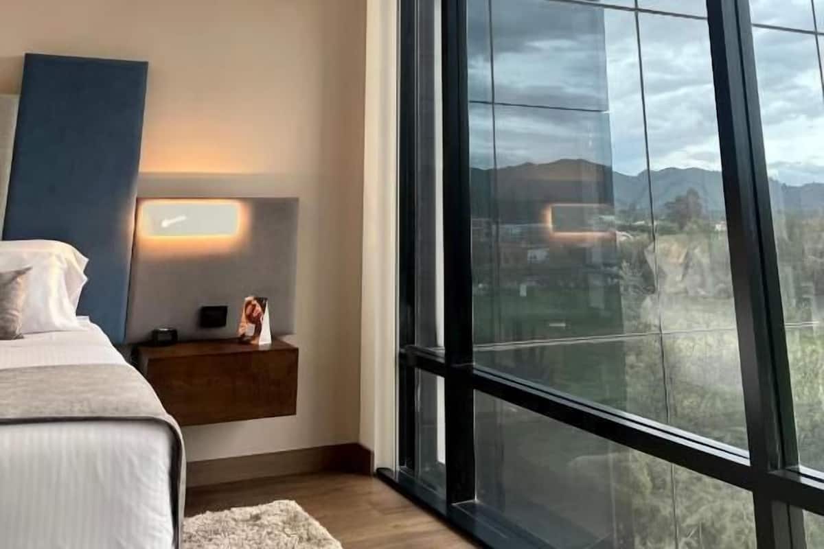 Un dormitorio con un ventanal grande, una cama, una mesita de noche y una lámpara de pared.
