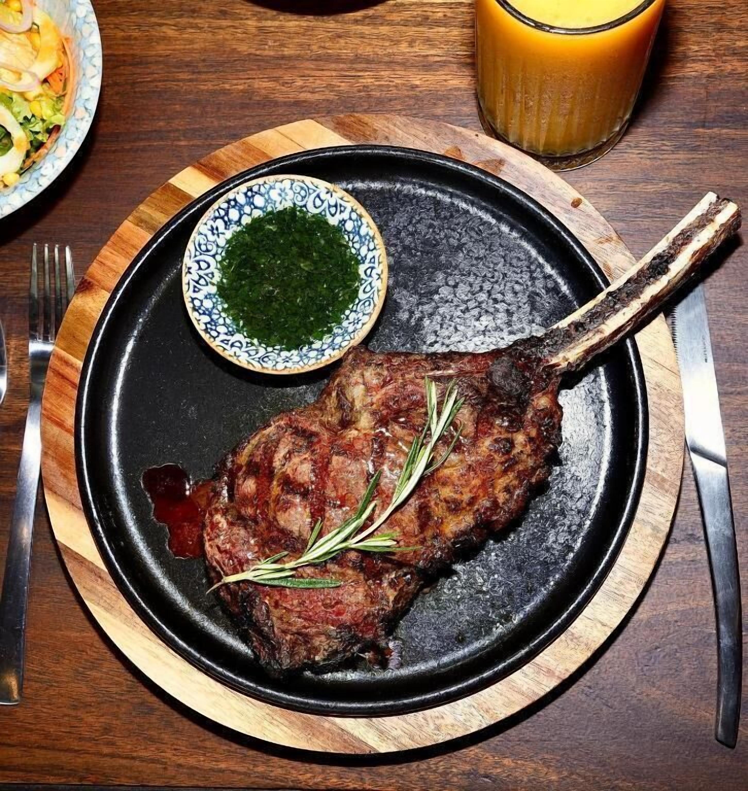 Een bord met een gegrilde steak, een klein schaaltje groensaus en een glas sinaasappelsap op een houten tafel.
