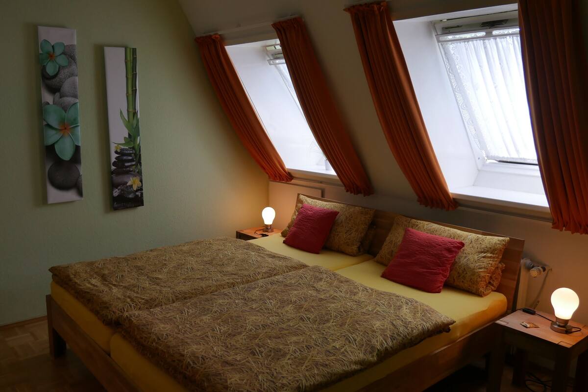 Un dormitorio con una cama, dos cuadros en la pared, una ventana con cortinas y una mesita de noche con lámpara.