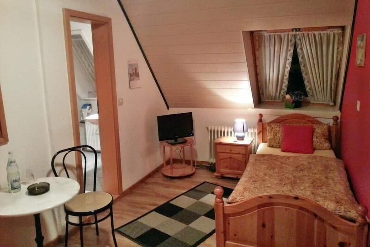 Un dormitorio con techo inclinado, una cama con cabecera de madera, una mesita de noche con lámpara, un televisor sobre un mueble bajo, una silla y una pequeña mesa con una botella y un posavasos para cenicero.