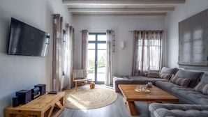 Een moderne woonkamer met een flatscreen televisie, een houten salontafel, een grijze bank en een groot raam met gordijnen.