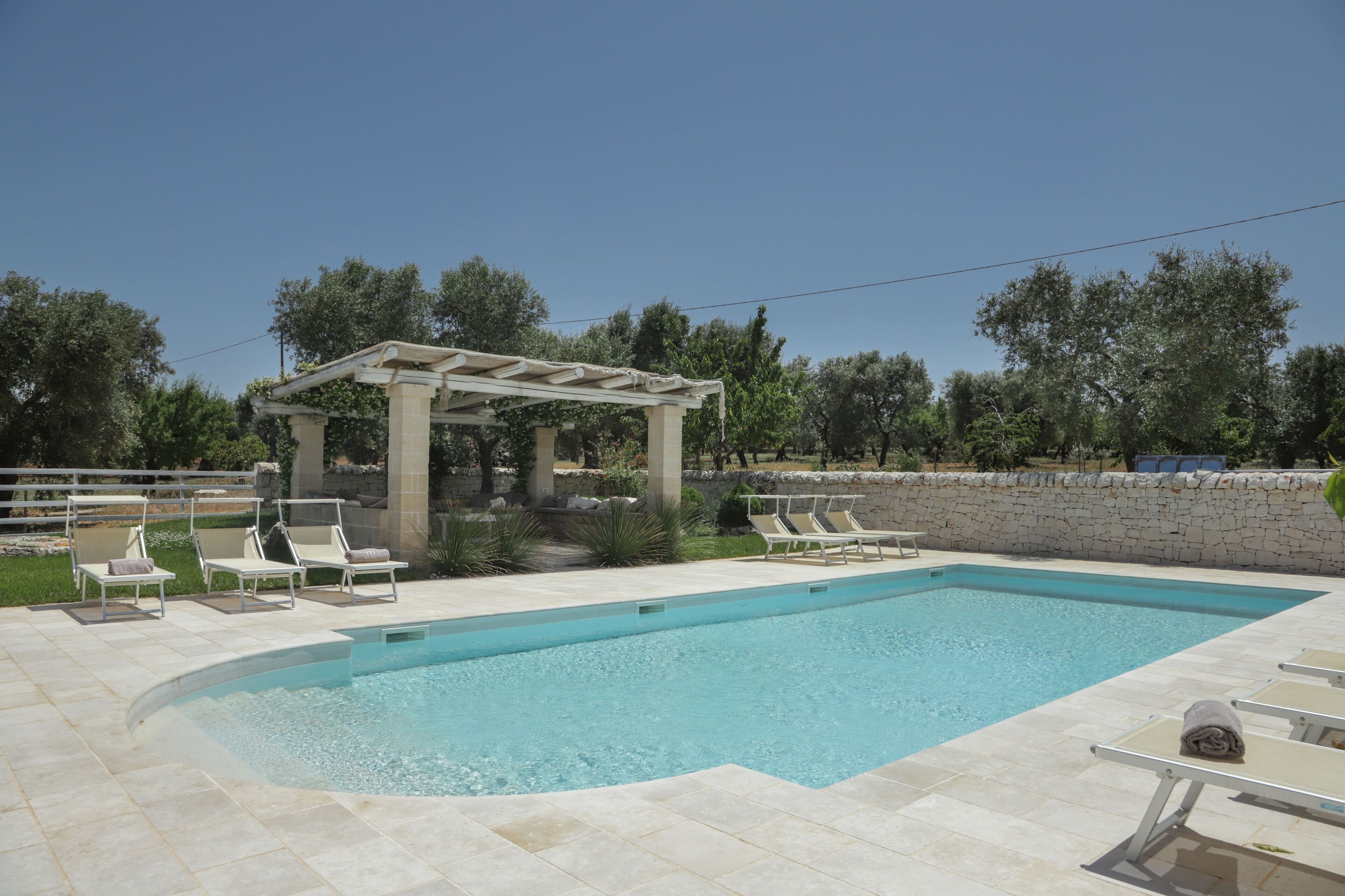 En rektangulär swimmingpool med en pergola och solstolar runt omkring.
