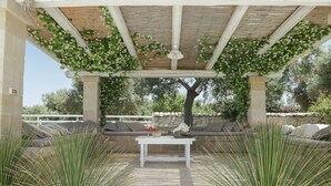 Ein möblierter Patio-Bereich im Freien mit weißem Tisch, gepolsterten Sitzmöglichkeiten und einer Pergola, die mit Kletterpflanzen begrünt ist.