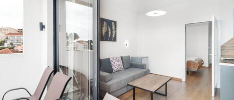 A modern apartment with a glass door leading to a balcony, a grey sofa, a wooden coffee table, and a bed in the adjacent room.