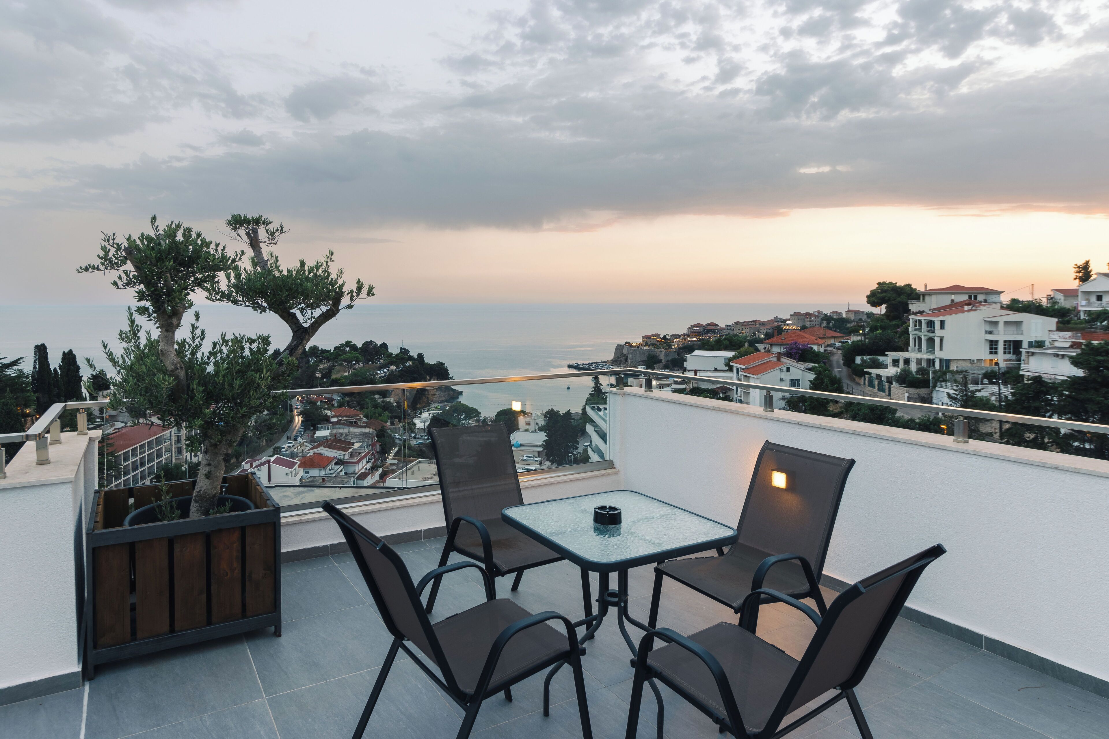 A rooftop terrace with a glass table and chairs, overlooking a coastal town and the sea.