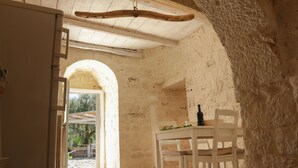 Um interior rústico de pedra com teto de madeira, uma mesa branca com cadeiras e uma garrafa sobre a mesa.