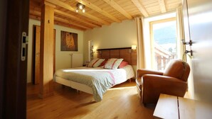 A bedroom with a wooden ceiling, a bed with patterned bedding, a chair, and a painting on the wall. - Le ValOmbre (Saint-Pierre-de-Chartreuse)