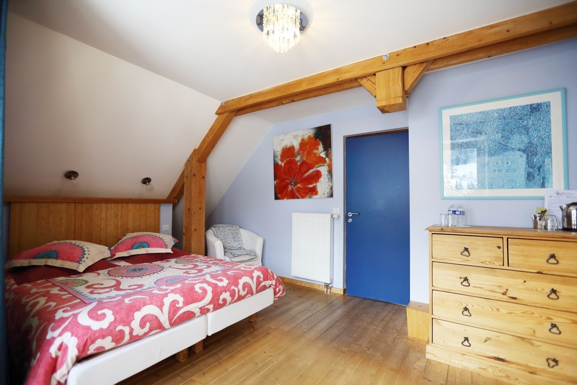 A bedroom with a sloped ceiling, wooden beams, a bed with patterned bedding, a chair, a dresser, and artwork on the walls.