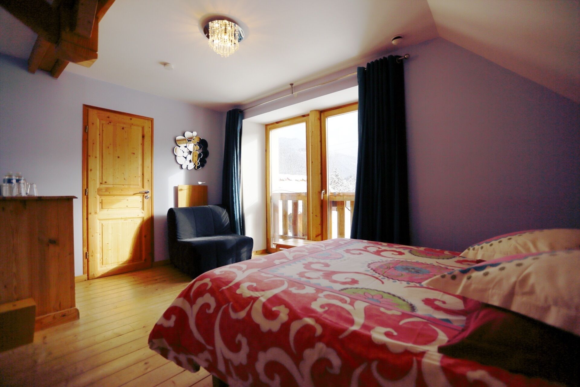 A bedroom with a wooden floor, a bed with a patterned bedspread, a chair, a wooden wardrobe, and a large window with a view of the outdoors.