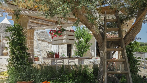 Un edificio de piedra con una pérgola de madera, plantas en macetas y una escalera.