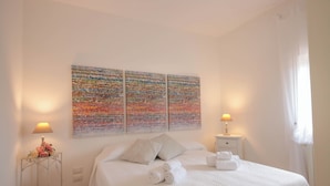 Una cama bien hecha con sábanas y almohadas blancas, una mesita de noche con lámpara, y un cuadro abstracto colorido sobre la cama.