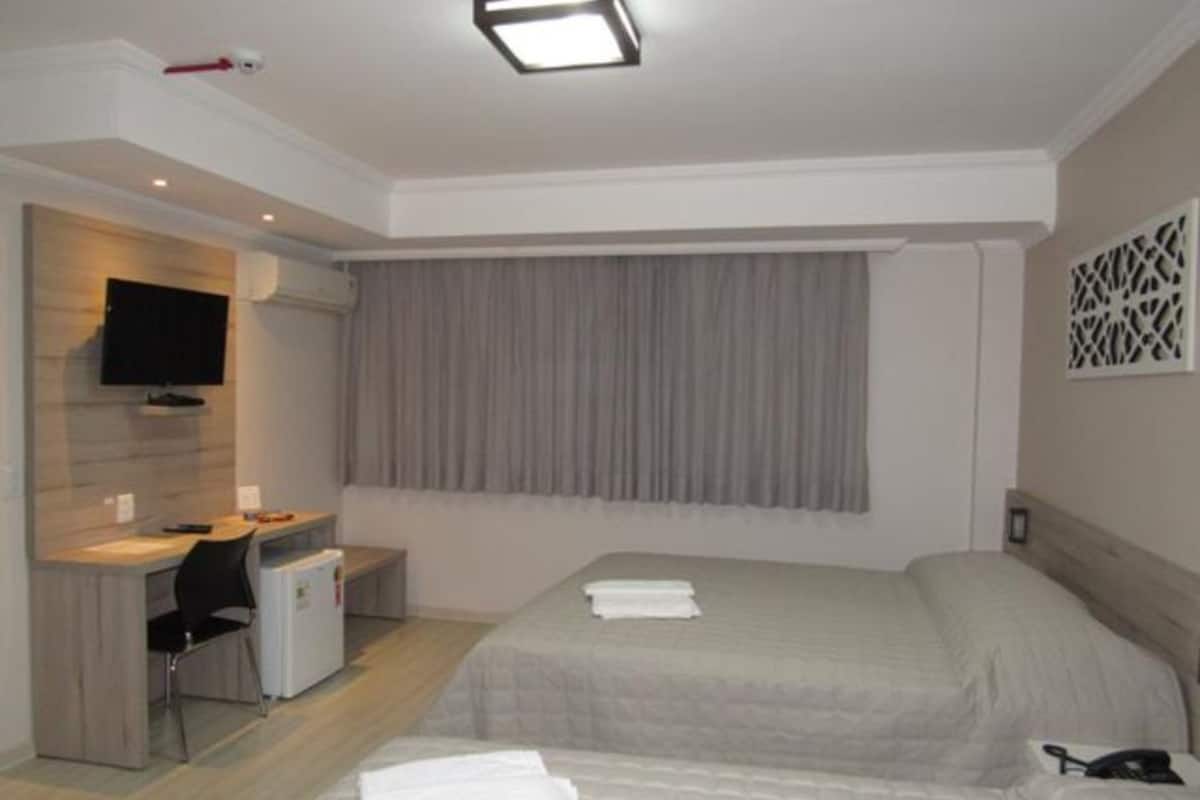 A hotel room with a bed, a desk with a chair, a television, and a window with curtains.