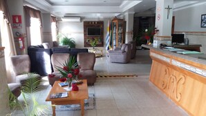 A hotel lobby with a reception desk, sofas, a coffee table with flowers, and a TV mounted on the wall. - Hotel Mariani (Lajeado)