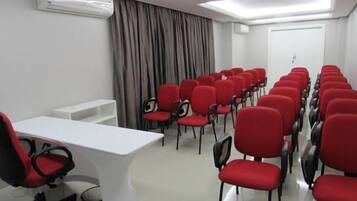 Uma sala de conferências com uma mesa branca, cadeiras vermelhas, um quadro branco e uma luminária no teto.