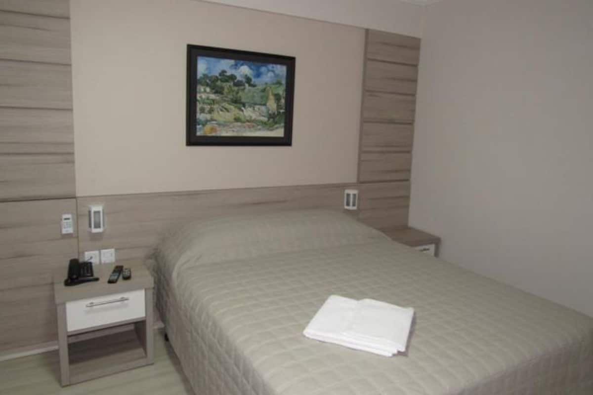 A neatly made bed with a folded white towel, a bedside table with a phone and a small lamp, and a framed painting on the wall.