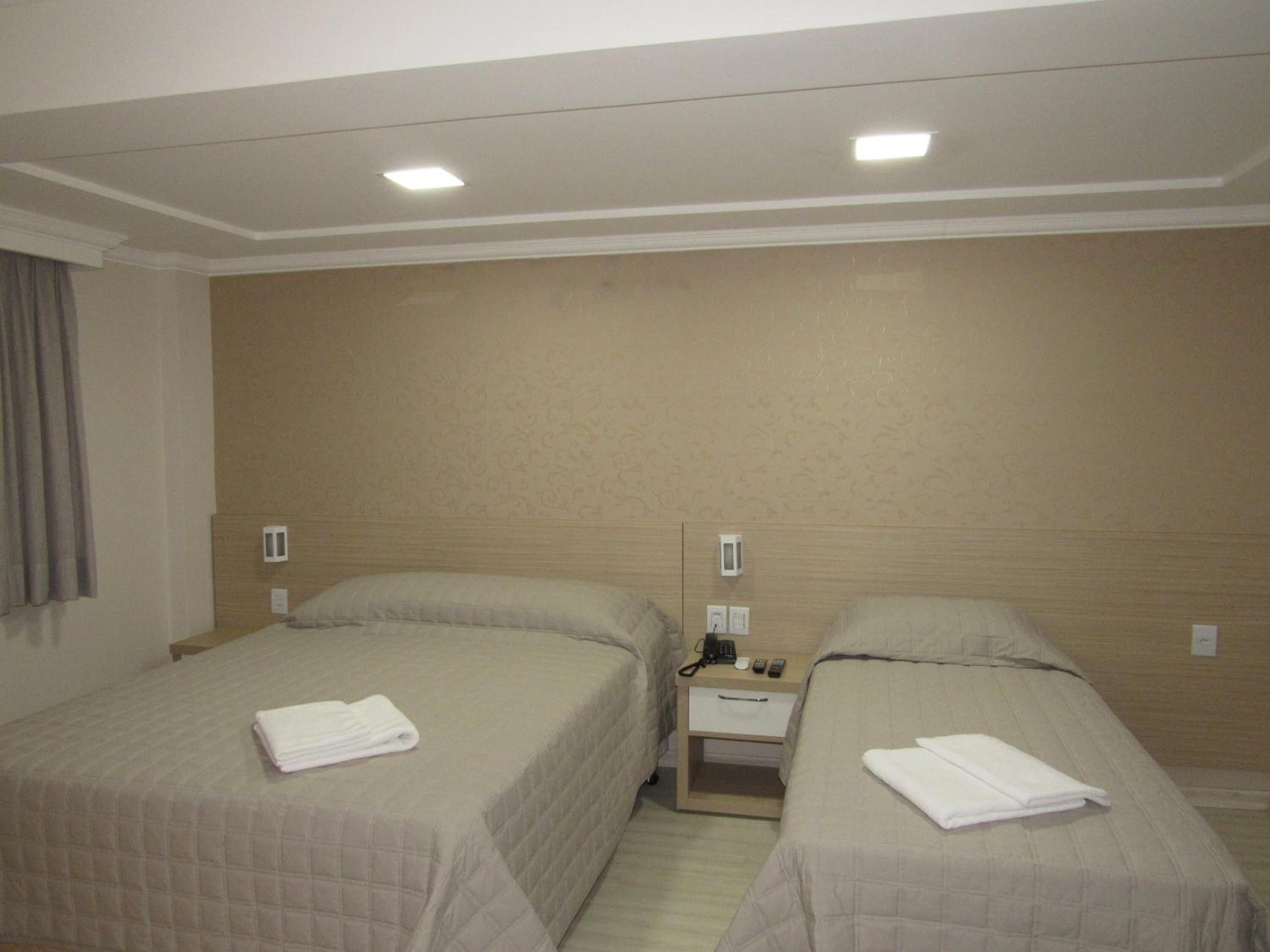 A hotel room with two beds, a bedside table with a phone and remote control, and a wall-mounted light fixture.