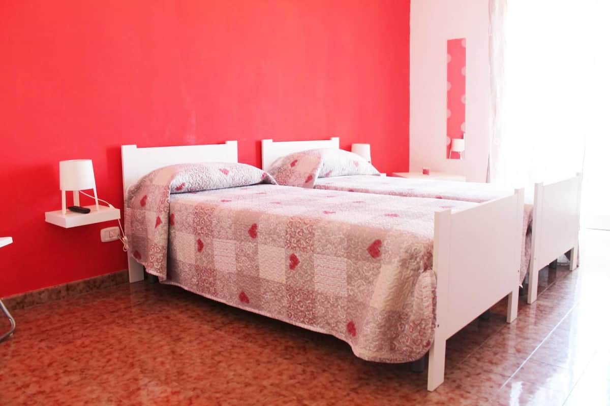A room with two beds, a red wall, a white bedside table with a lamp, and a tiled floor.
