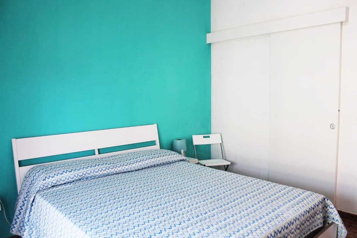 A bed with a patterned bedspread, a white headboard, a nightstand with a lamp, and a white wardrobe against a teal wall.